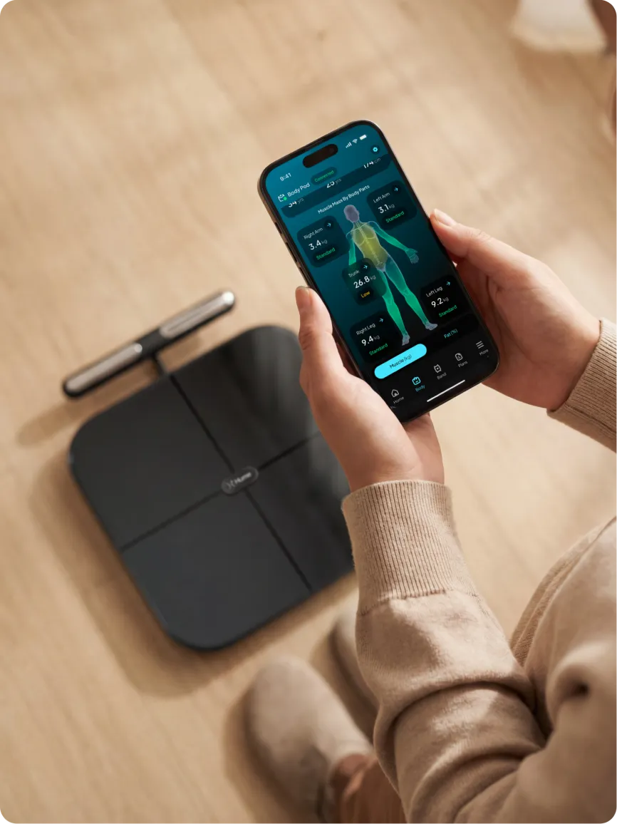 Person holding smartphone displaying Hume Body Pod app with full body composition analysis, with Hume Body Pod scale visible on the floor for connected health tracking.